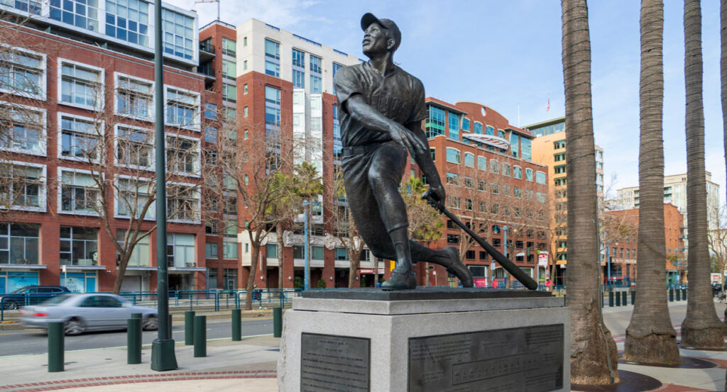 A statue of Willie Mays in front of Oracle Park with office buildings, restaurants and shops along the street and palm trees in San Francisco California USA